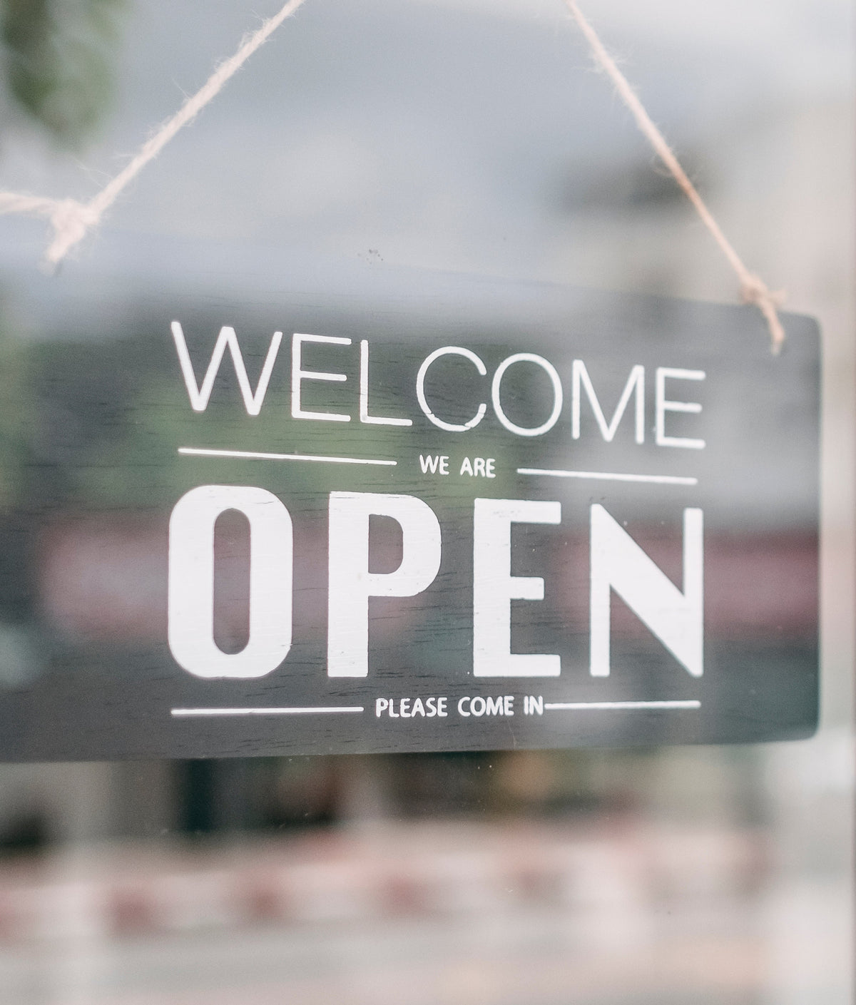 A rectangular black glass door sign with white text reading 'WELCOME WE ARE OPEN' hanging by a string on a storefront window next to a dark metal handle.